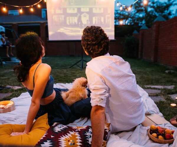 Two people with dog seated on grassy lawn with blankets and pillows watching movie on screen.